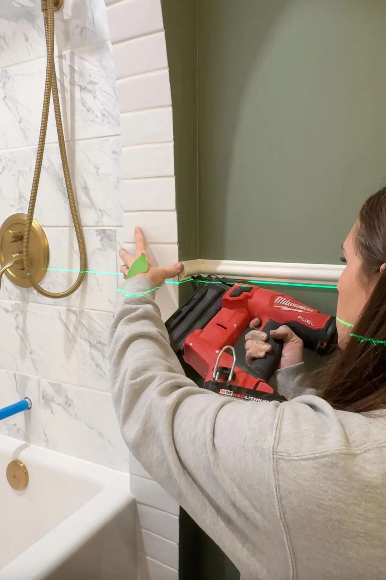 Close-up of a person using a red nail gun to secure chair rail molding near a white tile shower arch with brass hardware, demonstrating a bathroom trim installation.